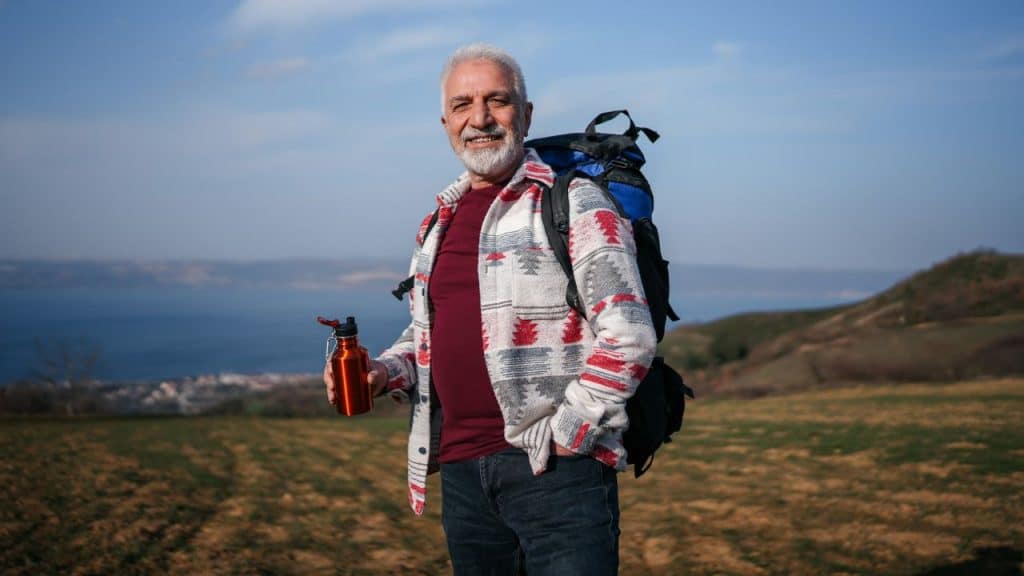 A smiling older man with a backpack holds a water bottle while hiking outdoors.
