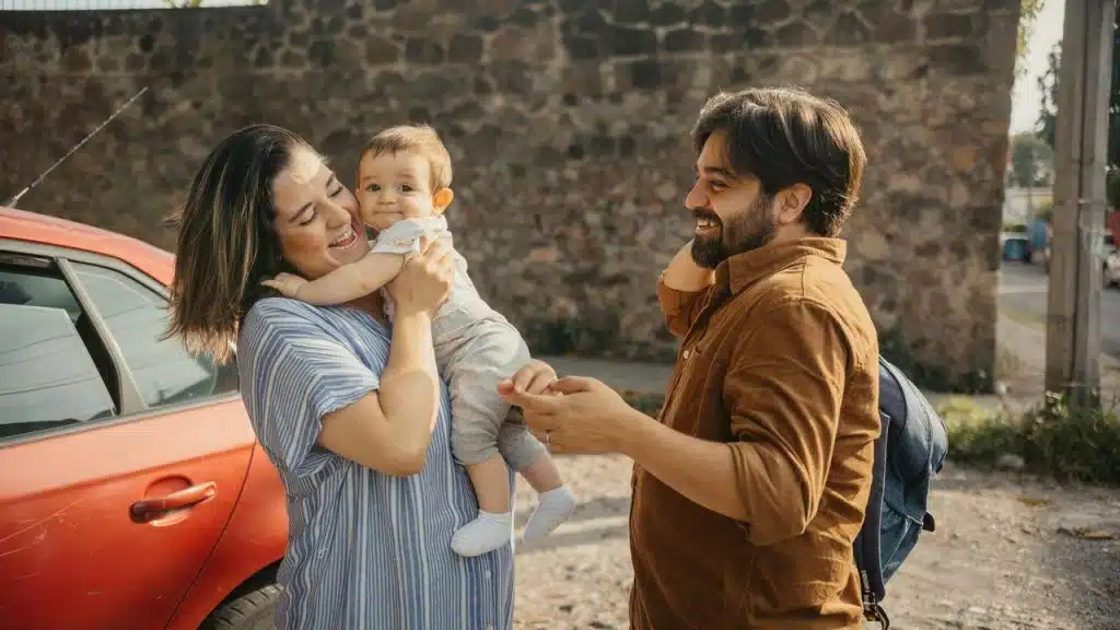 A smiling couple holding their baby while standing outdoors near a red car.