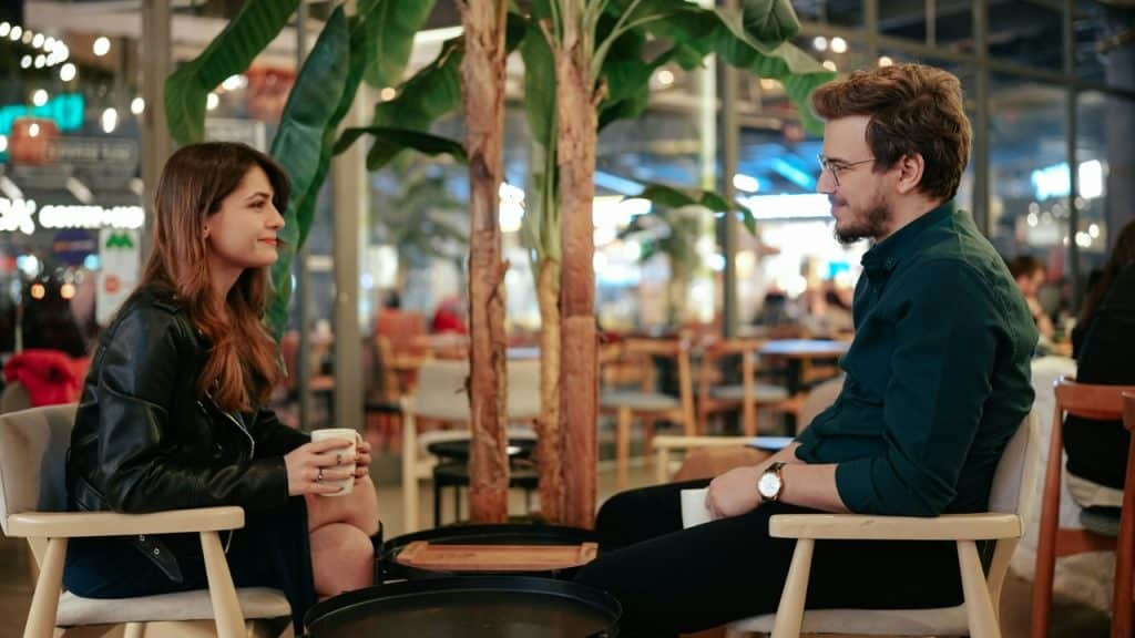 A man and woman sit in chairs at a cafe holding mugs and talking.