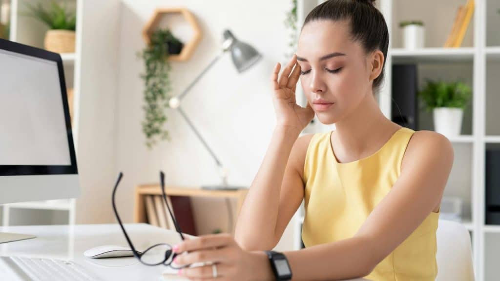 A woman in a yellow top looks tired while holding her glasses at a desk.
