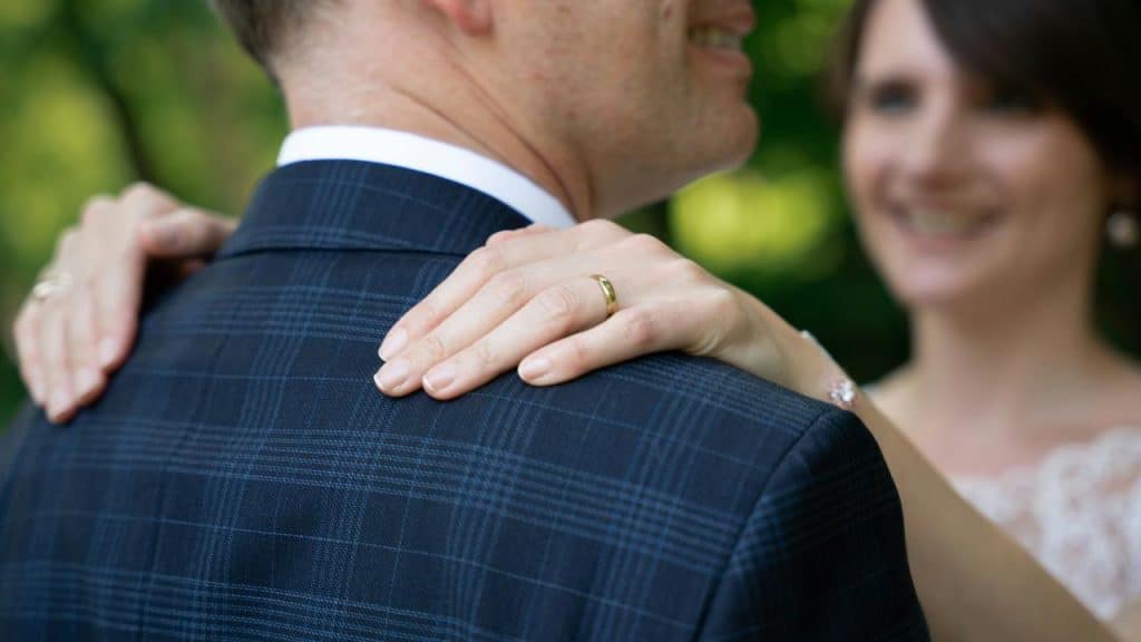 A bride’s ringed hands resting on a groom’s blue checked suit jacket.