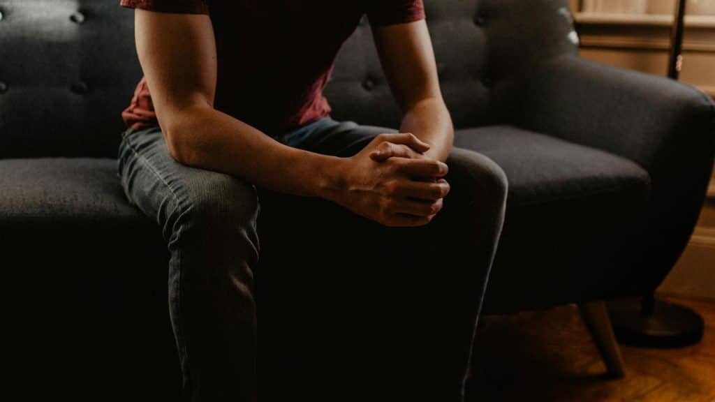 A person sitting on a couch with hands clasped in a dimly lit room.