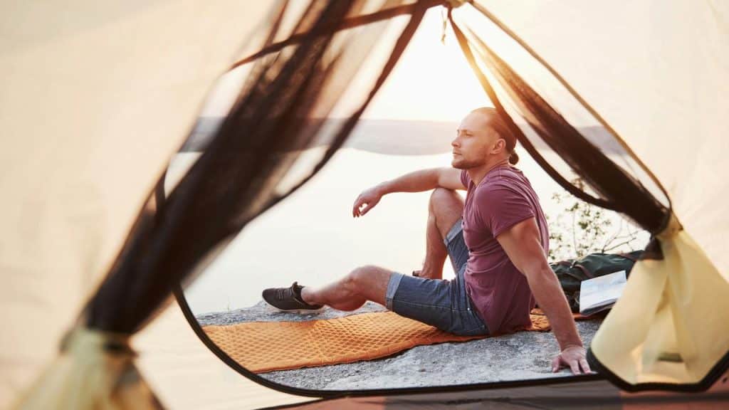 A man sitting at the opening of a tent, looking out over a calm body of water at sunset.