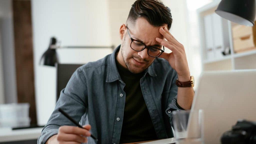 A frustrated man holding his head while working on a laptop.