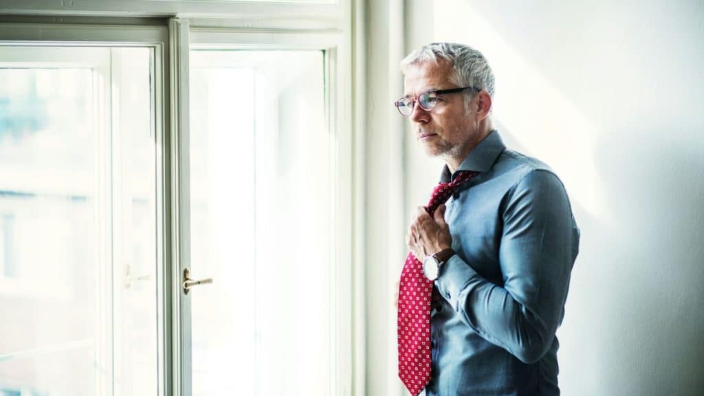 A man in a blue dress shirt and red tie looks out a bright window.