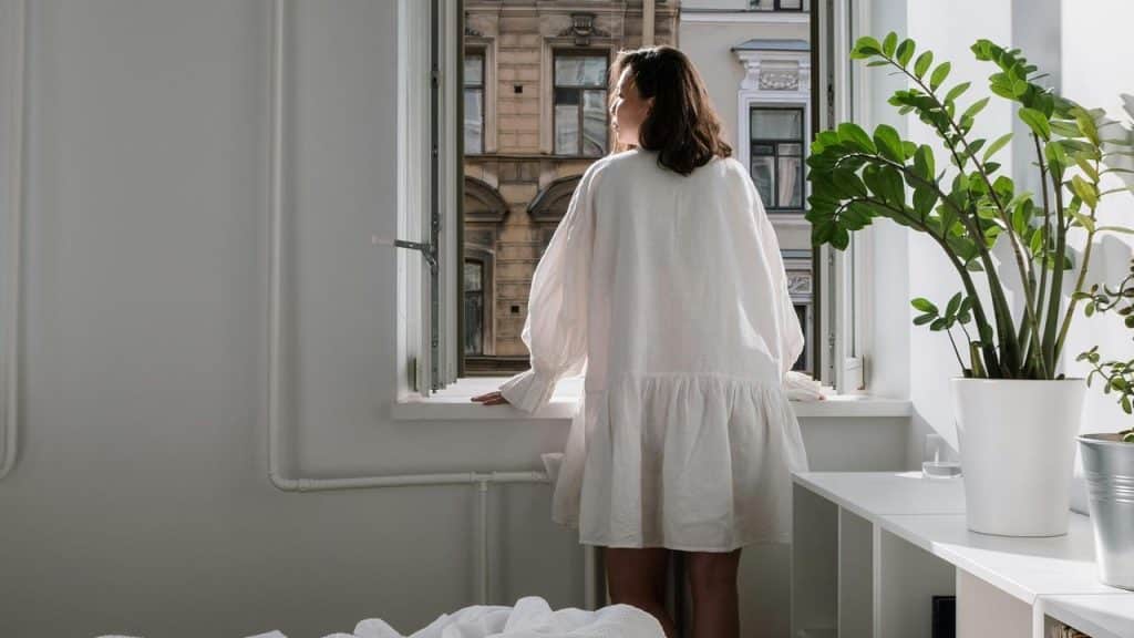 A person in a light dress standing by an open window and looking outside.