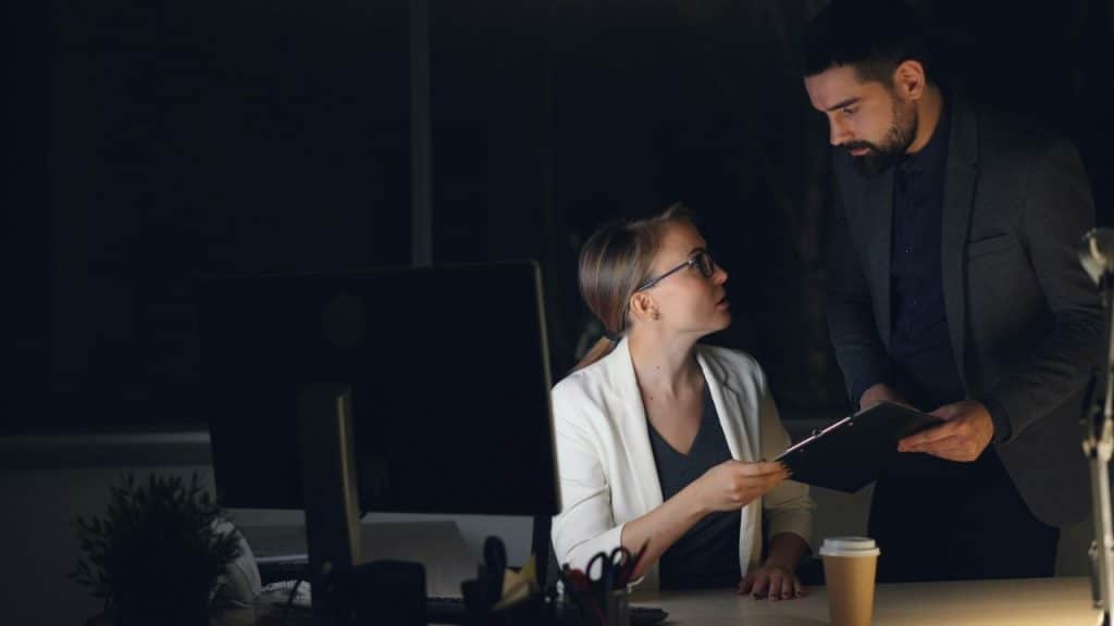 A woman and man in business attire look at a clipboard in a dark office.