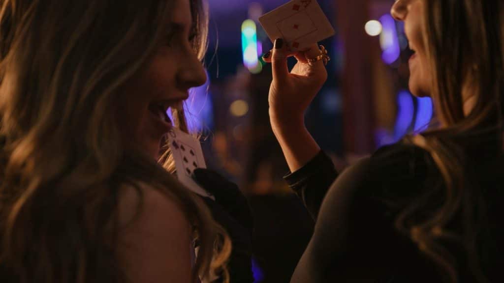 A pair of women laughing while holding playing cards in a dimly lit bar.