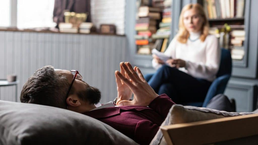 A man reclines on a couch, talking with a therapist holding notes.