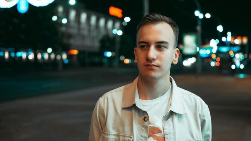 A man in a light jacket stands on a city street at night with lights blurred behind him.