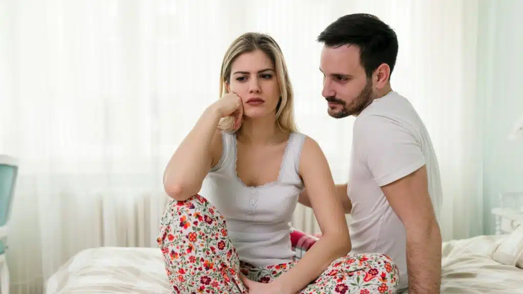 A man in a white shirt looks toward a woman sitting on a bed looking away.