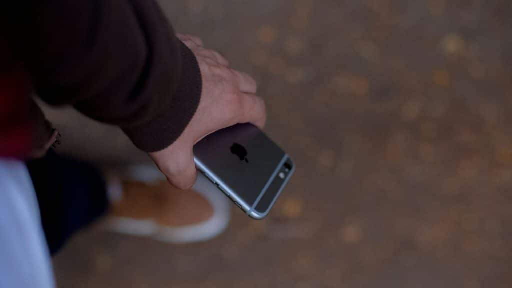 A hand holds a dark smartphone face-down while walking over a blurry, brown dirt surface.