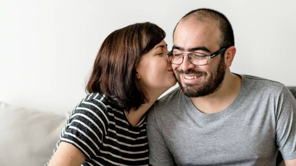 A woman in a striped shirt kisses the cheek of a smiling man wearing glasses.
