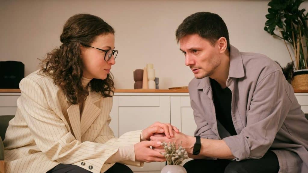A woman wearing glasses holds a man's hands while they look at each other indoors.