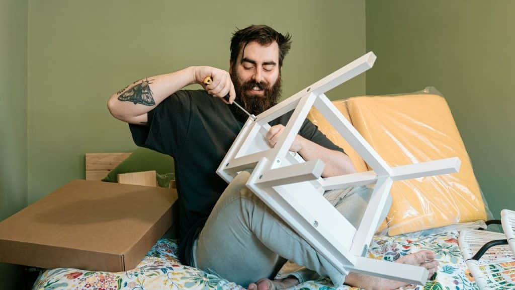 A bearded man with tattoos sits on a bed using a screwdriver to assemble a white chair.