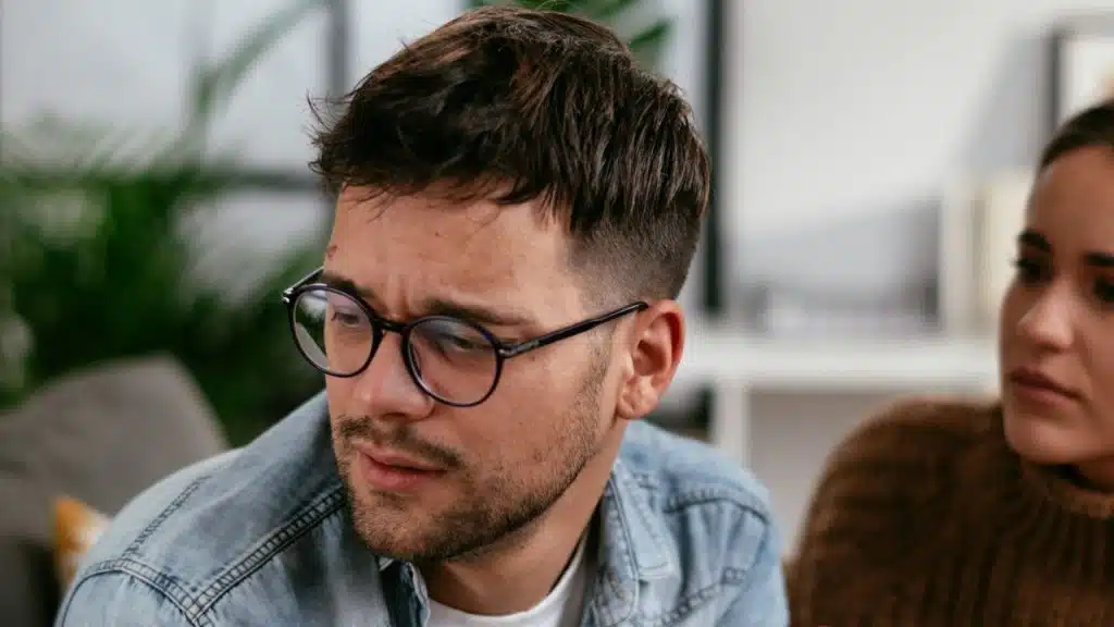 A man in a denim shirt looks away while a woman watches him from behind.
