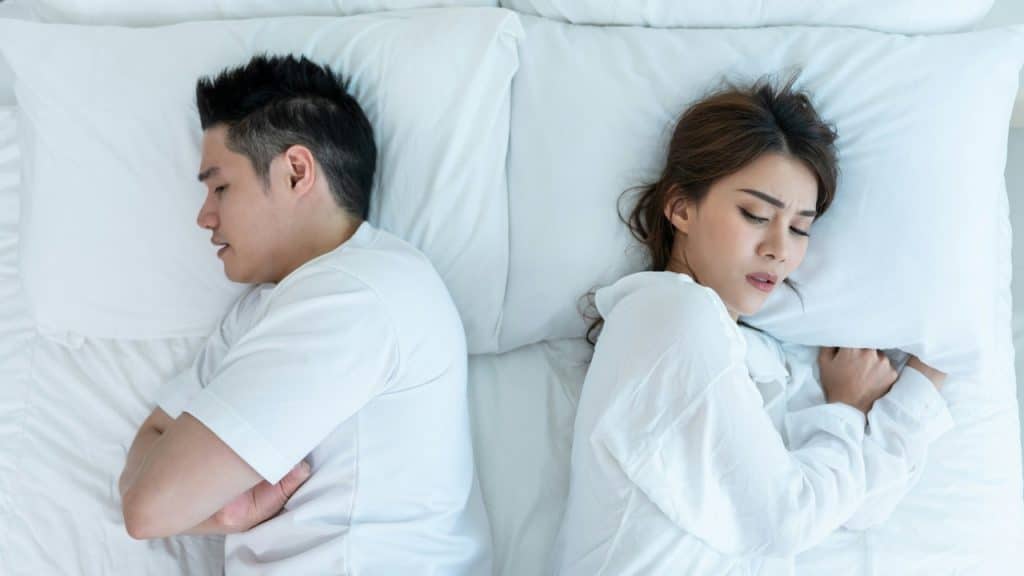 A man and woman lie in bed facing away from each other on white pillows.