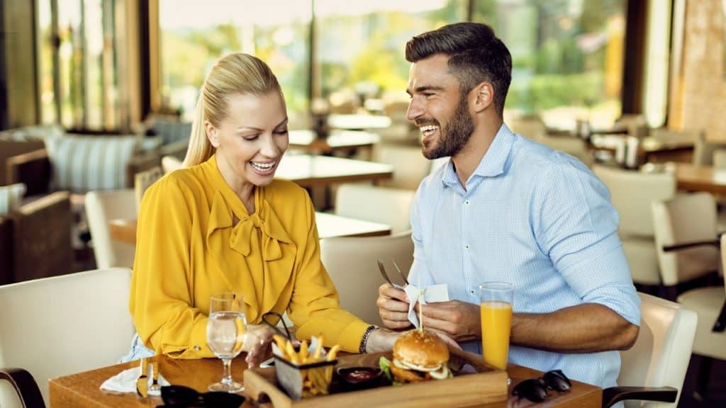 A man and woman sit at a restaurant table with food and drinks, smiling and laughing.
