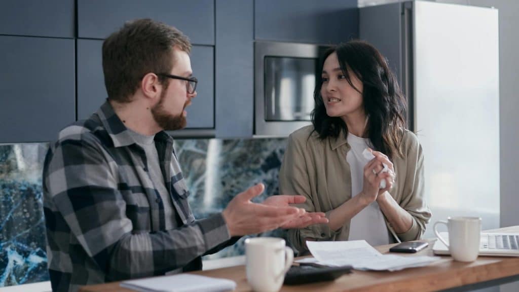 A man and woman sit at a kitchen counter with papers, talking and gesturing with hands.