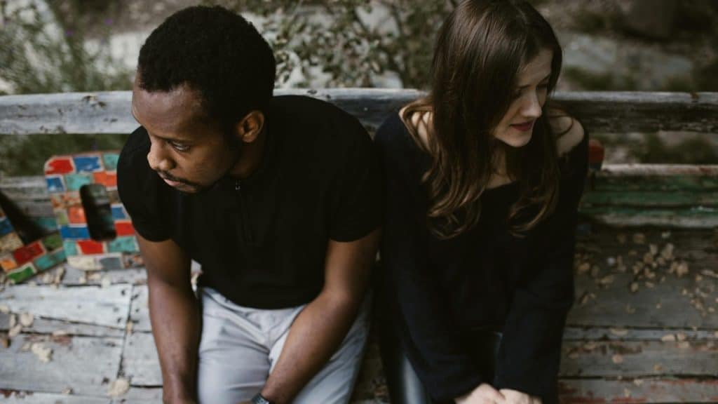 A man and woman sit back-to-back on a wooden bench, both looking in opposite directions.
