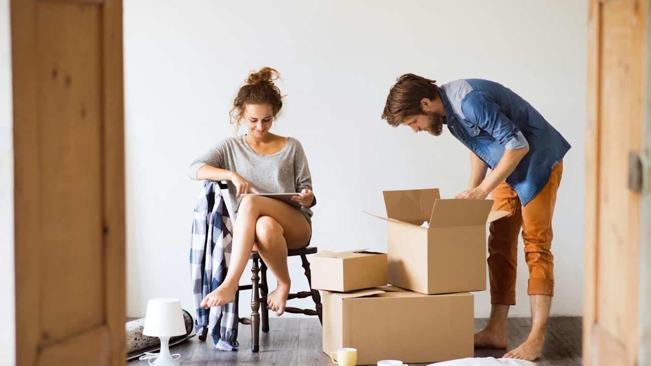 A woman sitting on a chair uses a tablet while a man stands over open cardboard boxes, both barefoot, in a bright room.