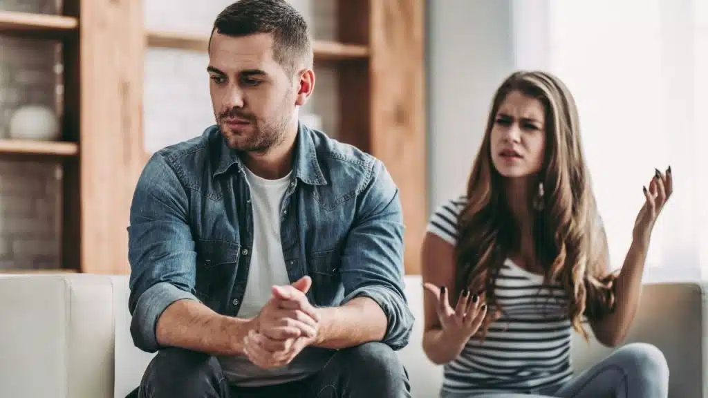 A man sits looking away while a woman in the background gestures with frustrated expressions.