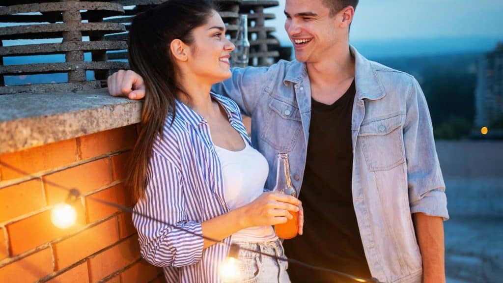 A smiling couple standing outdoors at dusk, chatting while holding a drink near string lights.