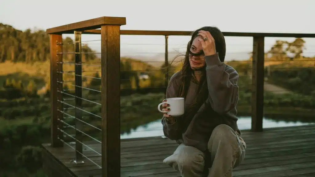 A woman holding a mug on a wooden deck overlooking a quiet landscape.