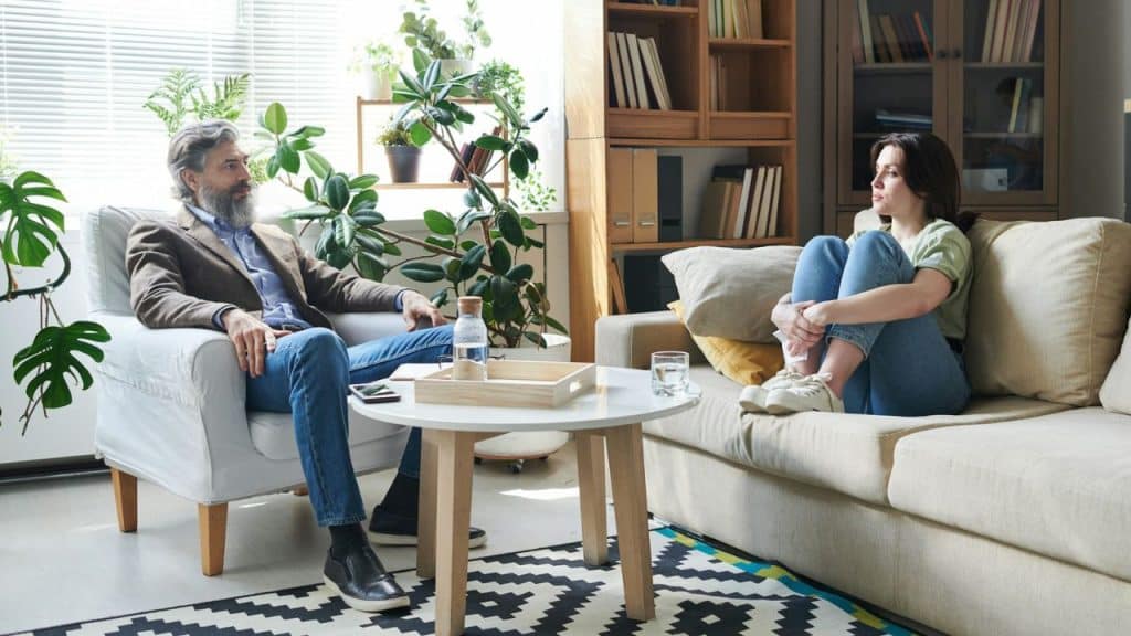 A man and woman sit apart in a bright living room, looking toward each other.