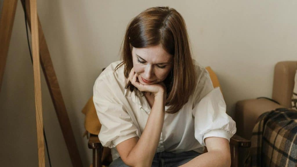 A woman in a white shirt sits with her chin resting on her hand, looking down.