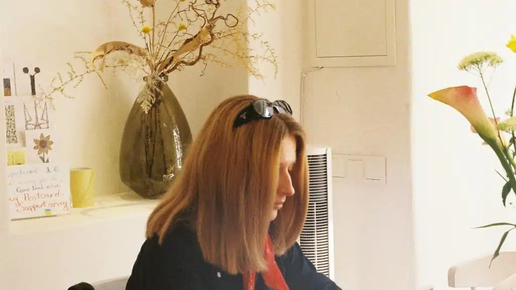 A woman indoors, looking down while seated near decorative vases and flowers.