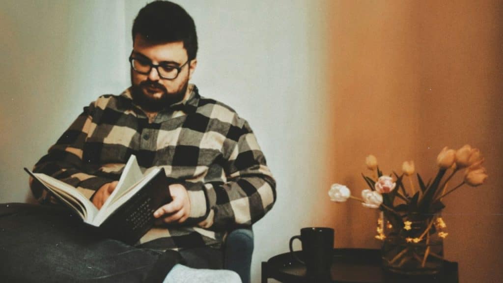A man with a beard and glasses wears a plaid shirt while reading a book indoors.
