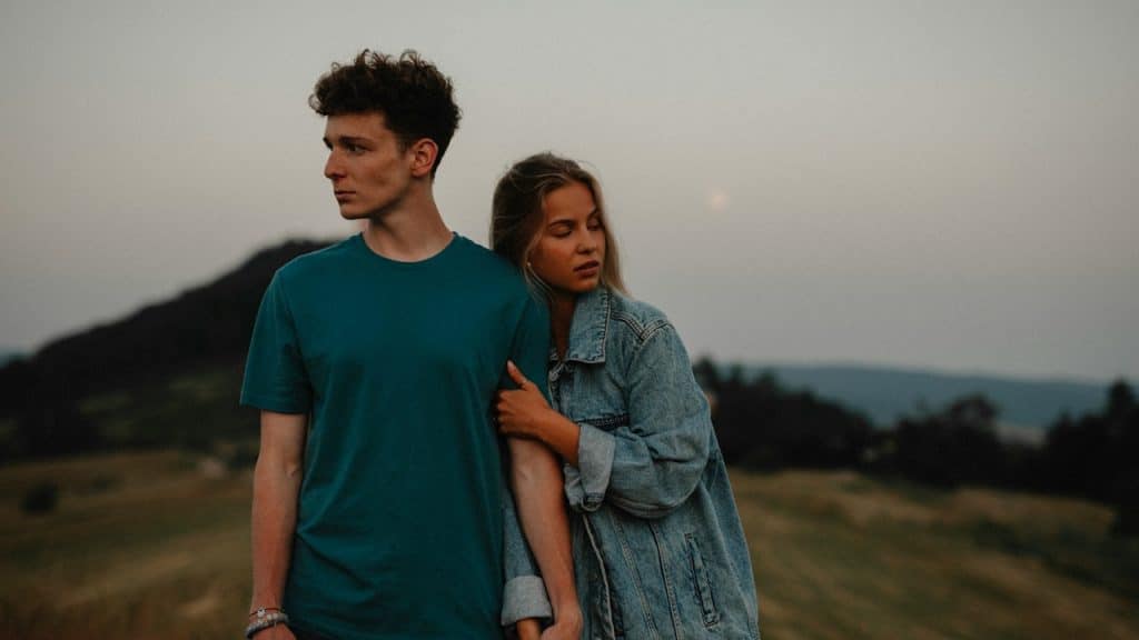 A young couple is standing outdoors in a field at dusk, holding hands, with the woman leaning her head on the man's arm.