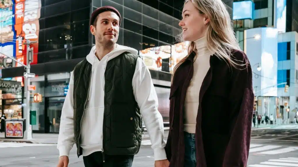 A man and woman hold hands while walking through a city street with tall buildings.