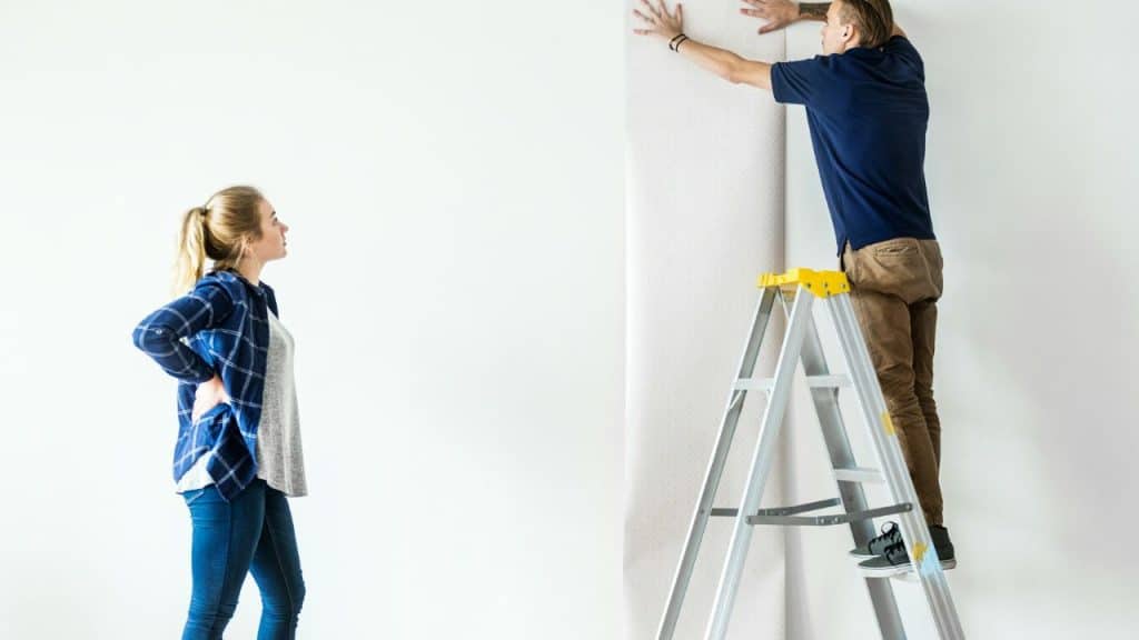 A woman watches a man standing on a ladder while he applies wallpaper to a wall.