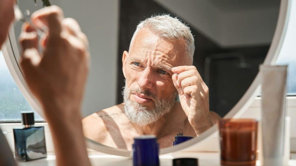 A grey-haired man looks into a round mirror while using tweezers on his eyebrow.
