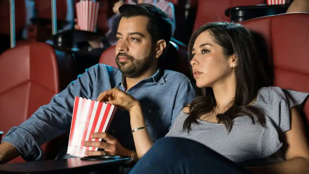 A man and woman sit in red theater seats, holding a striped popcorn bucket.