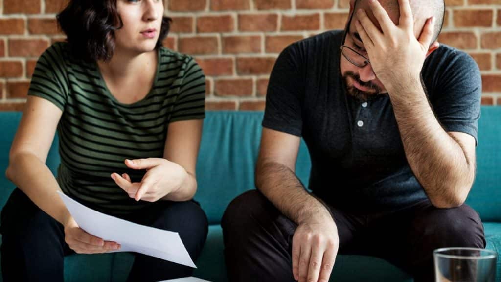 A woman holds a paper and gestures toward a man who is touching his forehead.
