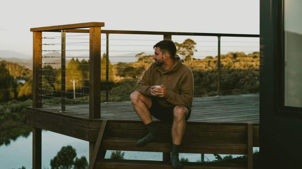 A man in a brown hoodie sits on wooden deck steps overlooking a lake and forest.