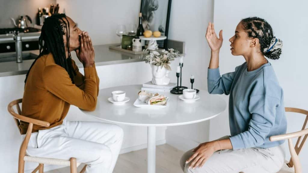 Two people sit at a white table with food, engaged in an expressive, serious conversation.