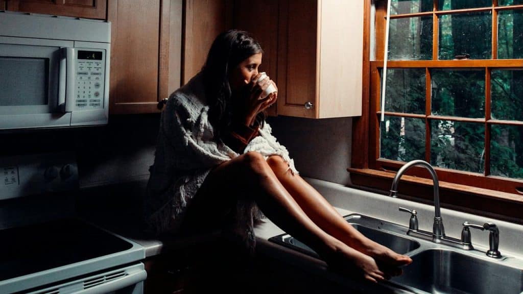 A woman sipping on a coffee cup while sitting on a kitchen counter and staring out of a window.