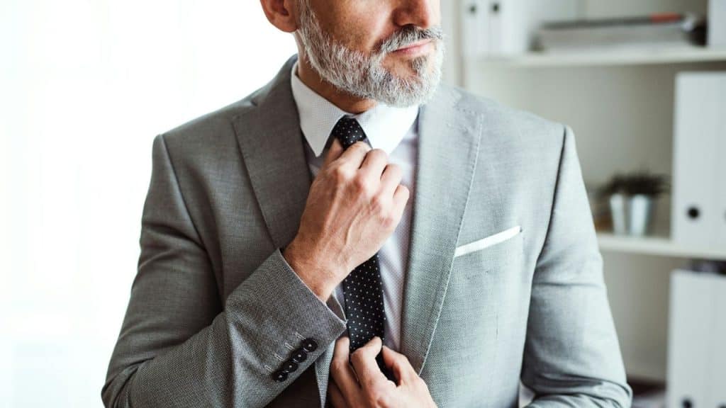 A grey-haired man in a suit adjusts his dark polka-dot tie with both hands.