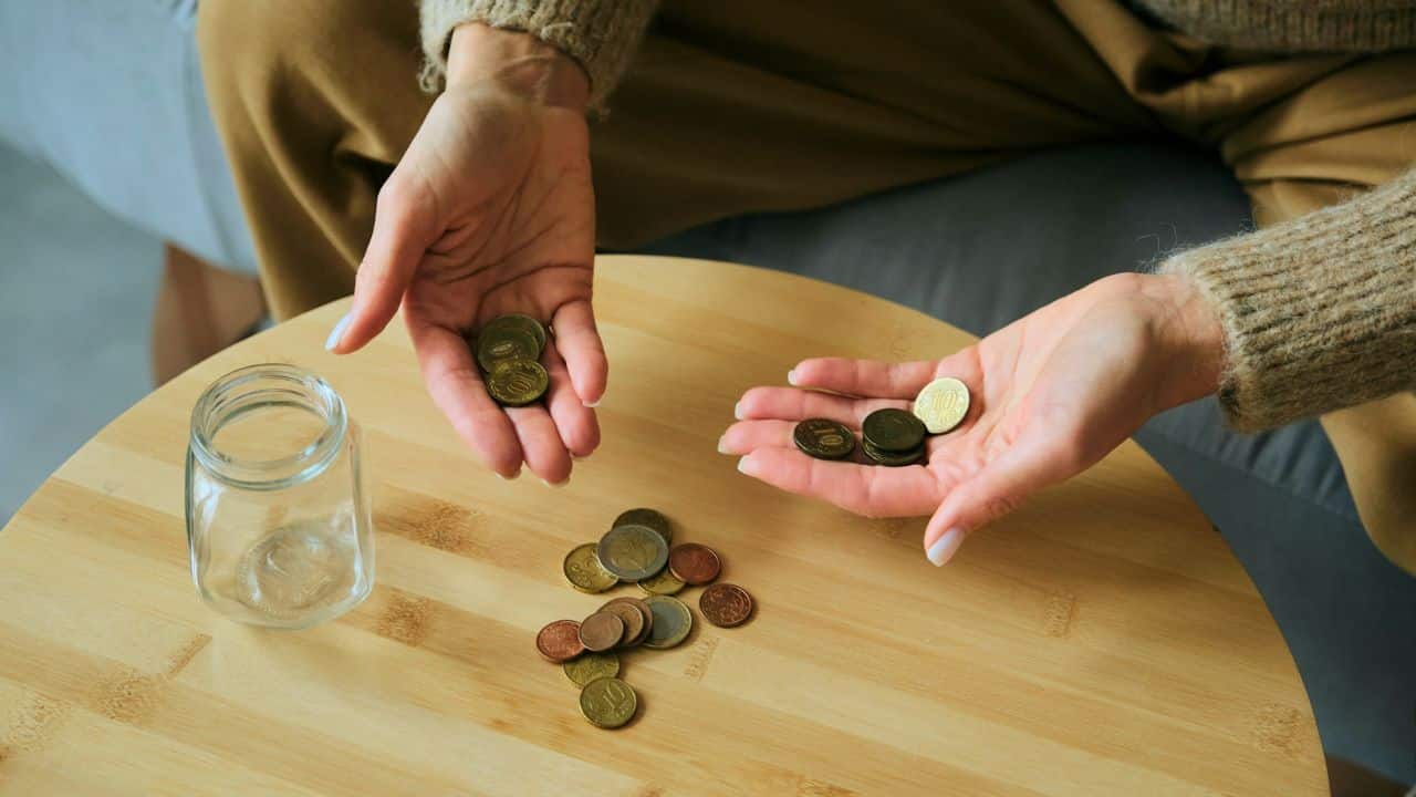 A person holds various coins in their open palms over a wooden table and glass jar.