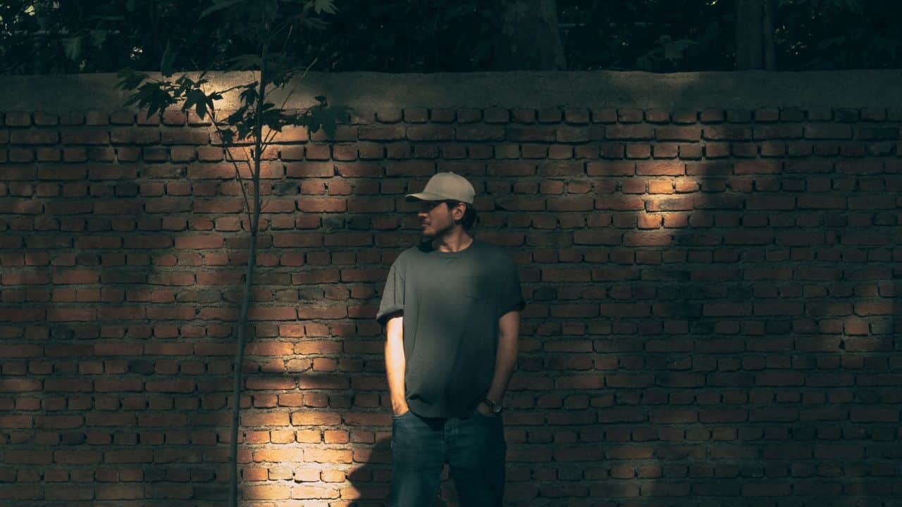 A man in a baseball cap and grey t-shirt stands against a brick wall outside.