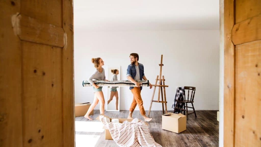 A couple carrying a rolled rug while unpacking boxes in a bright room.