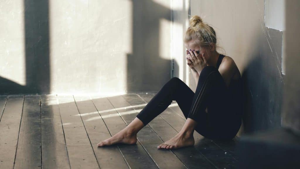 A woman sitting on the floor against a wall, covering her face with her hands.