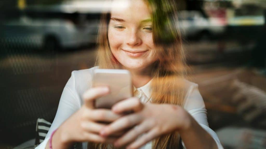 A smiling young woman looking at her smartphone through a window reflection.
