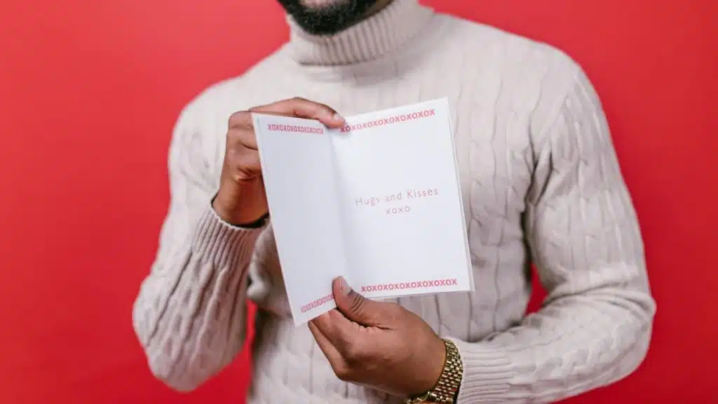 A man in a cream cable-knit sweater holds open a greeting card against a red background.