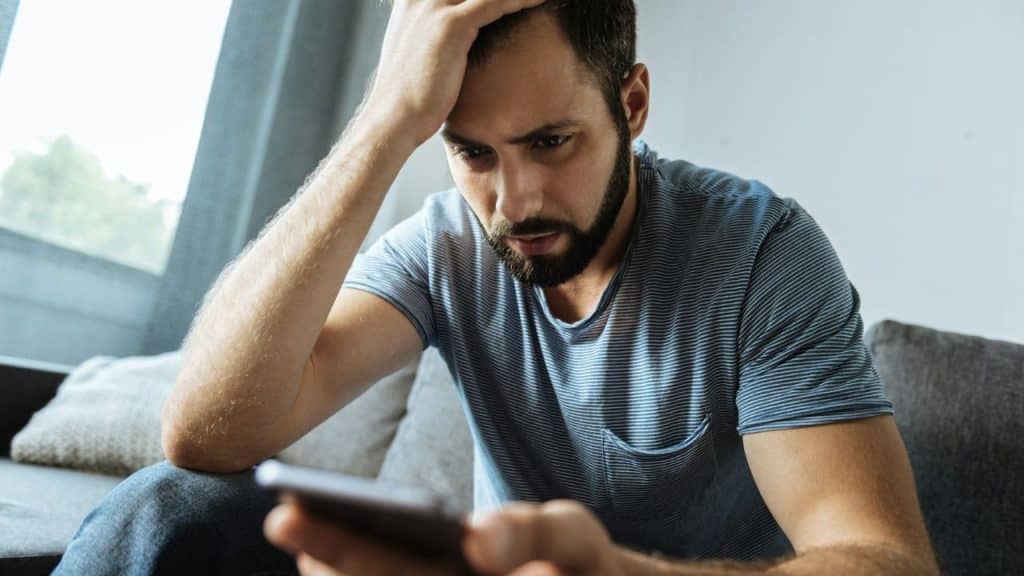 A bearded man in a striped shirt holds his forehead while looking at a smartphone.
