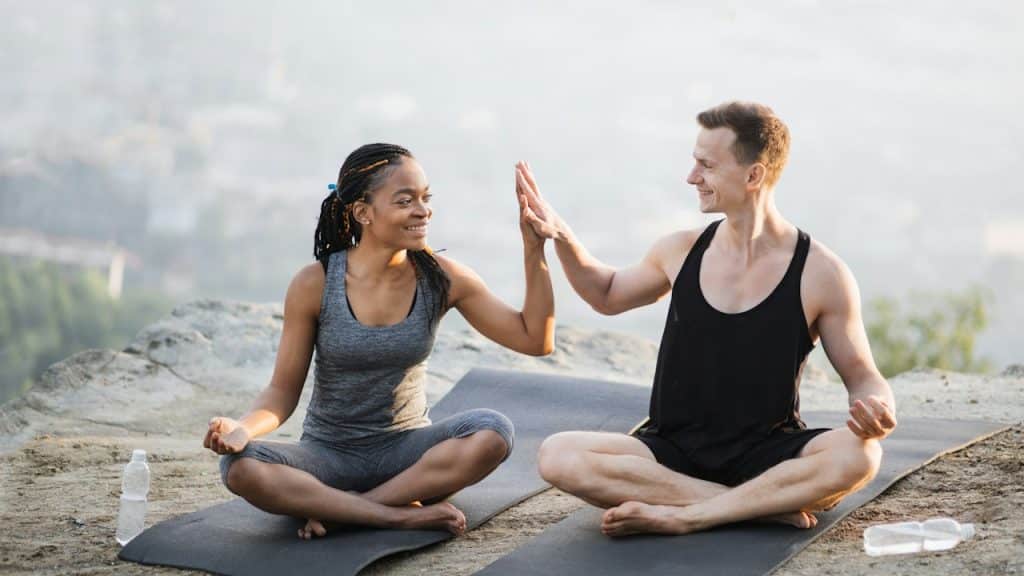 A couple high-fiving each other while sitting cross-legged on yoga mats on top of a cliff.